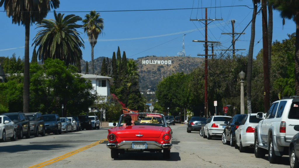Czerwone auto kabriolet na ulicy, w tle znak Hollywood w Los Angeles. Dookoła palmy oraz samochody.
